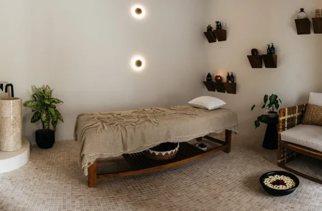 A serene spa room features a massage table, a stone sink, potted plants, and wooden shelves with products, all set against cream walls and a tiled floor.