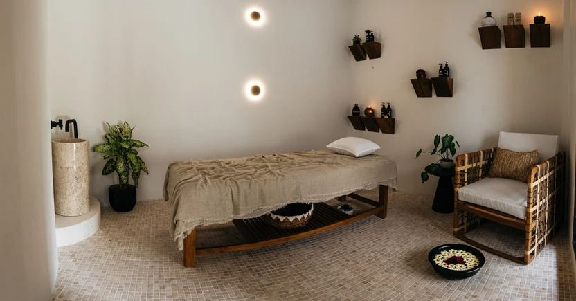 A serene spa room features a massage table, a stone sink, potted plants, and wooden shelves with products, all set against cream walls and a tiled floor.