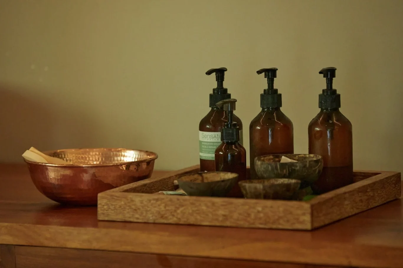 A hammered copper bowl and a wooden tray with amber pump bottles and small coconut shell bowls are arranged on a dark wooden surface.