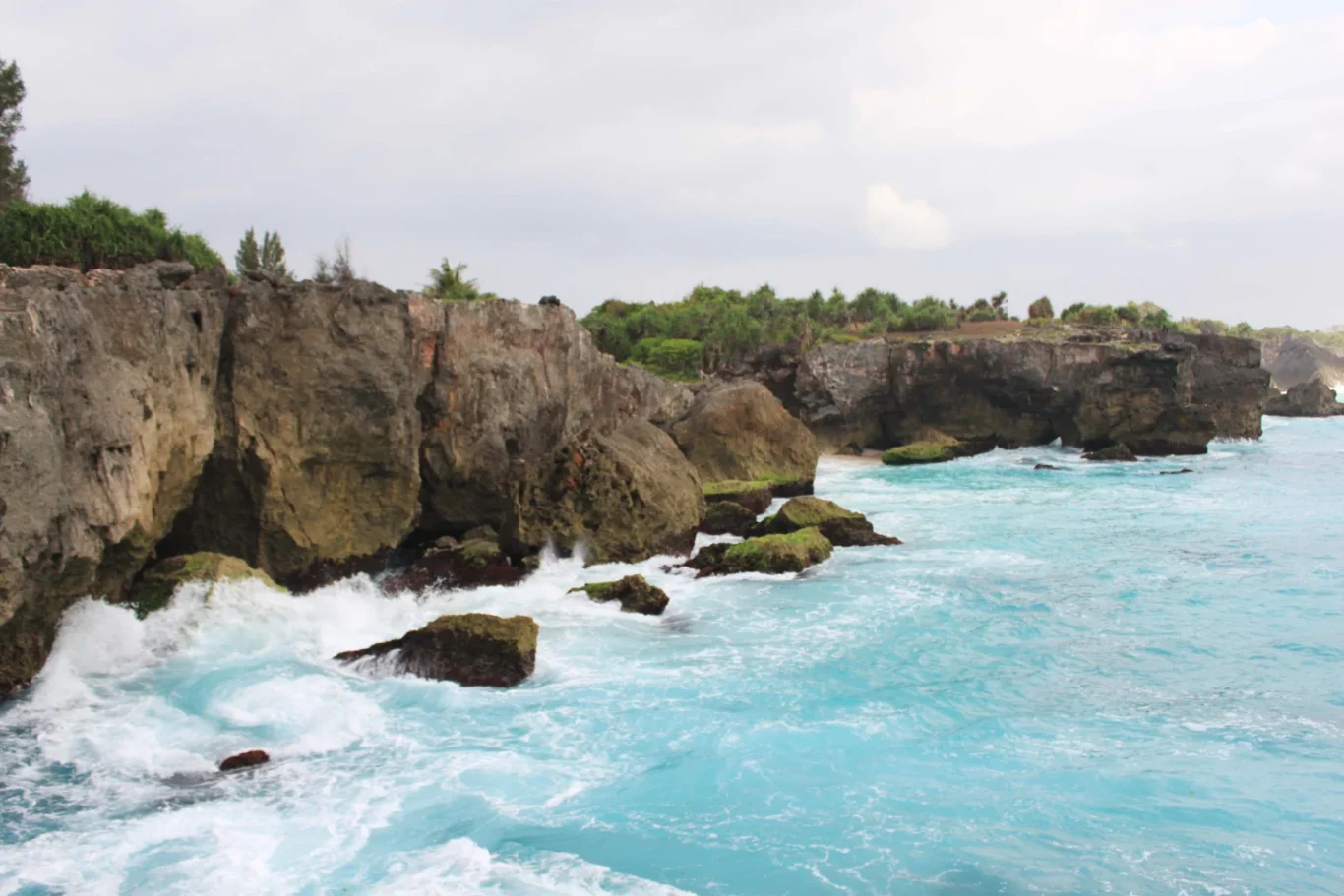 Rugged grey-brown cliffs topped with green foliage meet a vibrant turquoise ocean where white waves crash against rocks under an overcast sky.