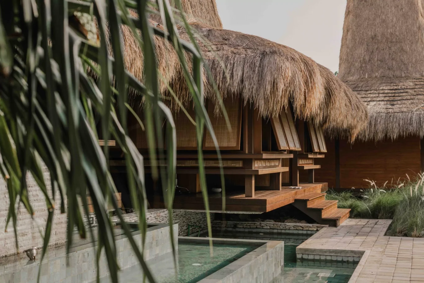 Traditional thatched-roof bungalows with wooden platforms and steps stand beside light blue water features, framed by blurred green palm leaves and tall grasses.