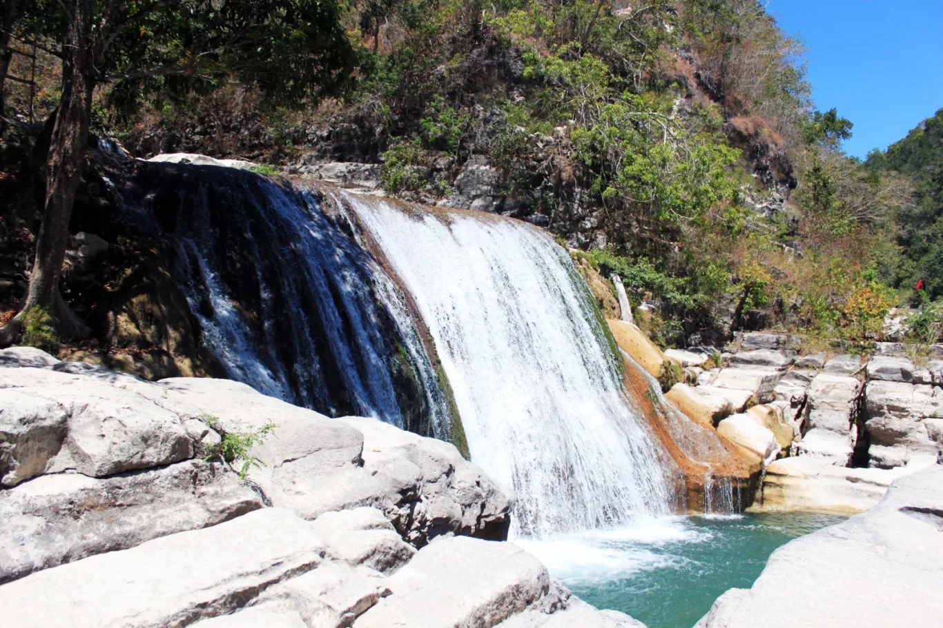 A wide waterfall cascades over dark and light brown rocks into a turquoise pool, surrounded by lush and dry green foliage under a clear blue sky.