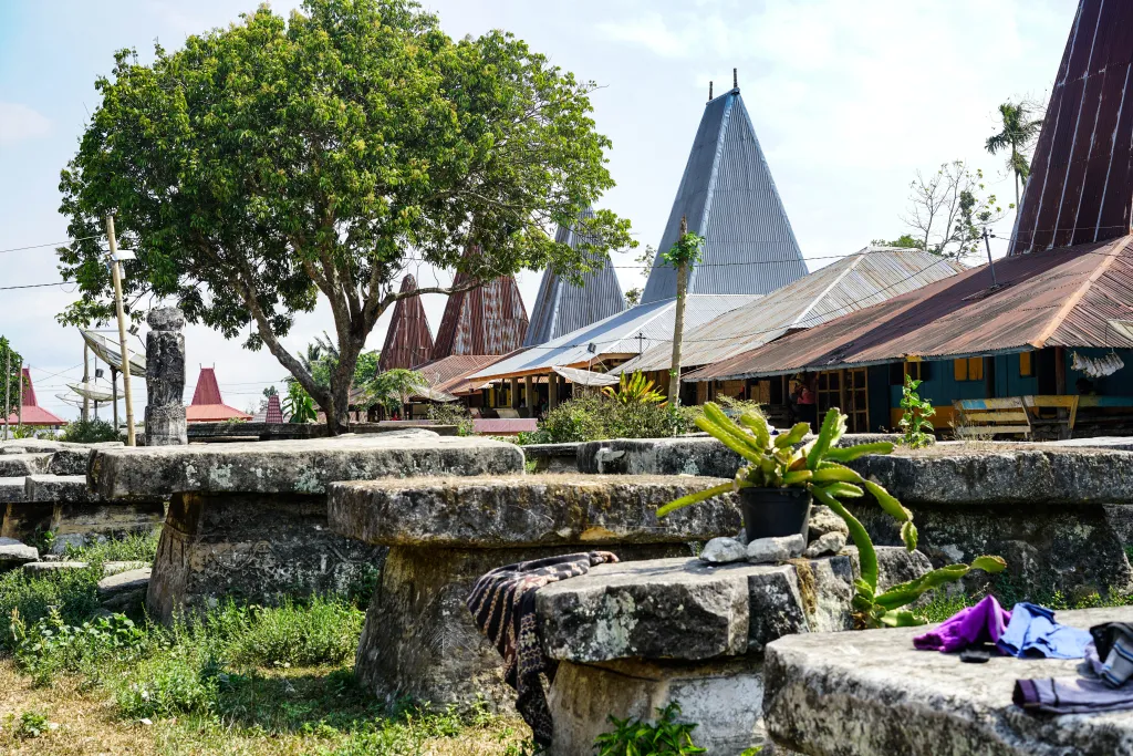 Large grey stone megalithic tombs sit in the foreground of a traditional village with tall, conical-roofed houses and a large green tree under a bright sky.