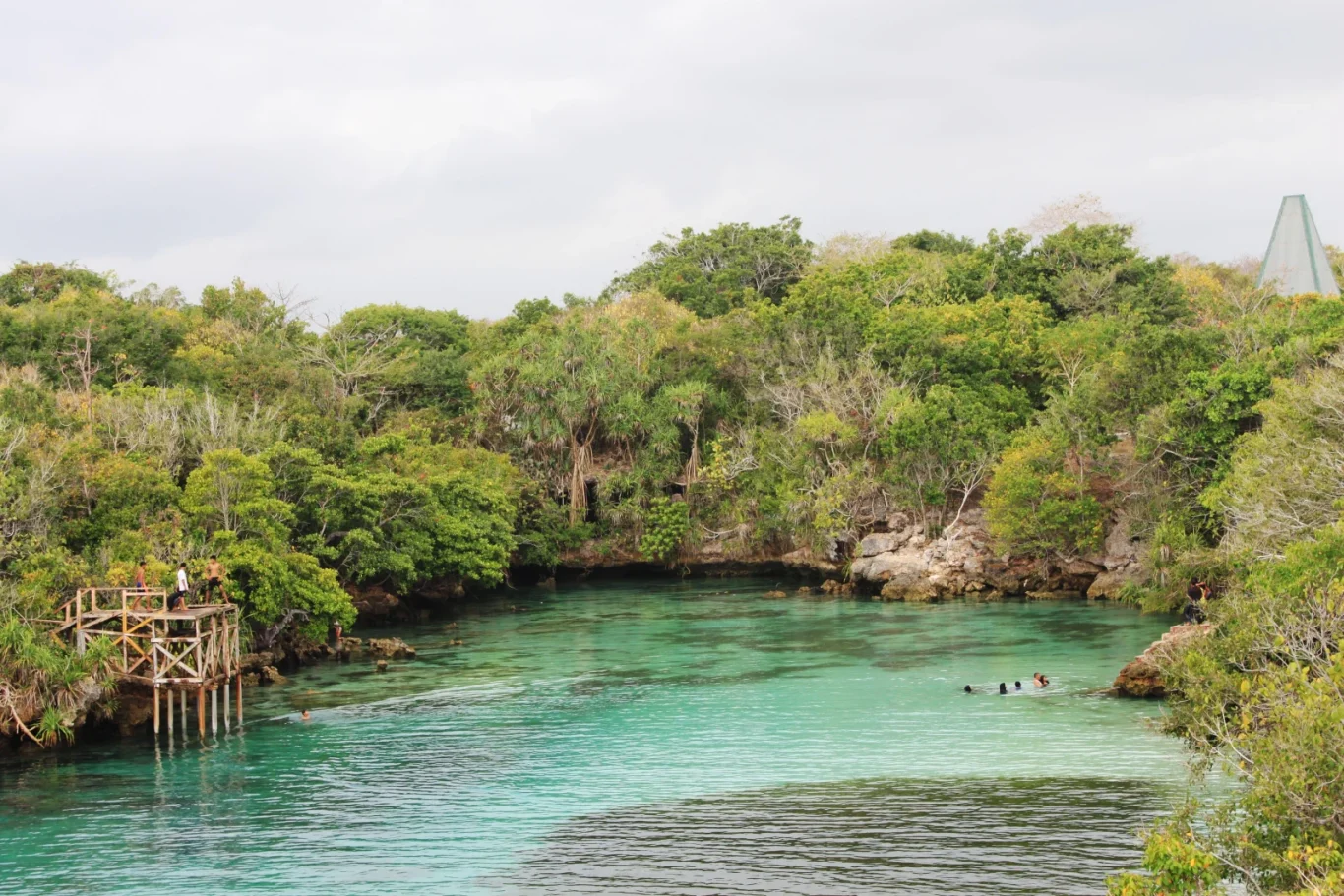 A clear turquoise lagoon is surrounded by lush green tropical trees under a cloudy sky, with a wooden platform on the left and people swimming.