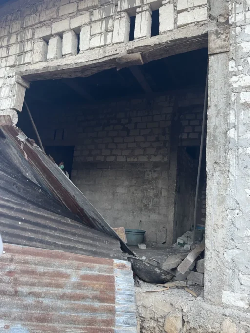 A partially collapsed building entrance with corrugated metal sheets in the foreground, revealing a dark interior where a person in a mask is visible.