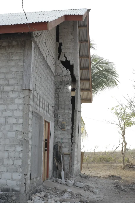 A light gray concrete block building with a large vertical crack on its side and a corrugated metal roof stands in a dry, dusty landscape.