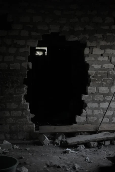 A dark, rough brick wall with a large, irregular opening reveals a shadowy interior, while debris and wooden planks lie on the dirt floor.