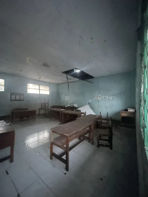 A dimly lit, empty classroom with light blue walls, a grey tiled floor, and several dark wooden desks and chairs. A large hole in the ceiling lets in bright light, and two windows are visible on the left.