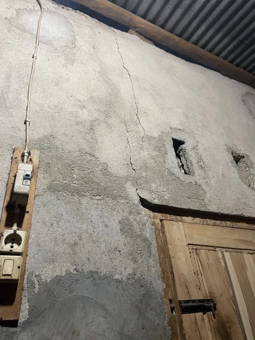 A cracked, light gray concrete wall features a wooden plank with electrical outlets and a circuit breaker, next to a rustic wooden door under a corrugated metal roof.