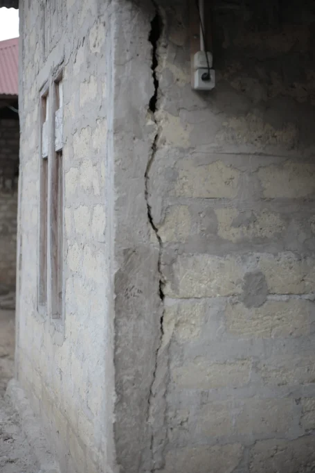 A close-up of a building corner with a large vertical crack, featuring rough light gray and yellow concrete blocks, a window frame, and an electrical outlet.
