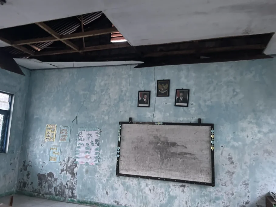 A dilapidated classroom interior features a large hole in the ceiling revealing wooden beams, peeling light blue walls, a crooked whiteboard, and framed portraits.