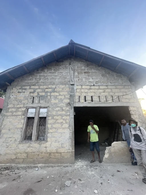 A light-colored brick building with a corrugated metal roof stands under a clear blue sky. Three men are near its entrance; one smokes, another leans, and a third wears a mask.