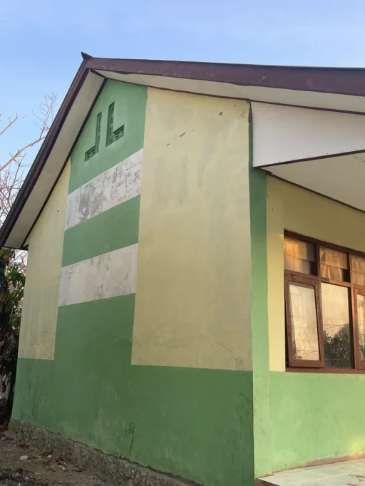 A light green and yellowish building with a white stripe and brown-framed windows stands under a clear blue sky, showing some weathered paint.