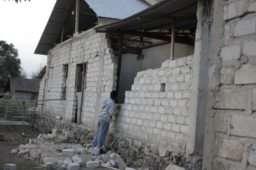 A person in a grey hoodie and blue jeans leans against a partially built light brick wall, looking into an opening, with debris on the ground and another building in the background.