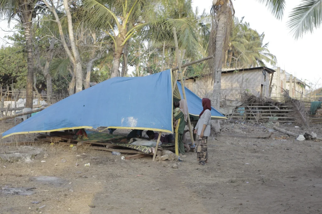 A blue tarp shelter stands on dry, dusty ground with several people, including women and children, gathered around it under palm trees and sparse vegetation.
