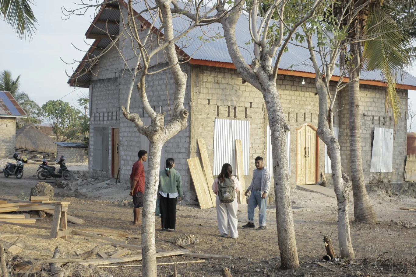 Four people stand in a dusty yard with two large trees in the foreground, facing a grey brick house with a metal roof and wooden doors.