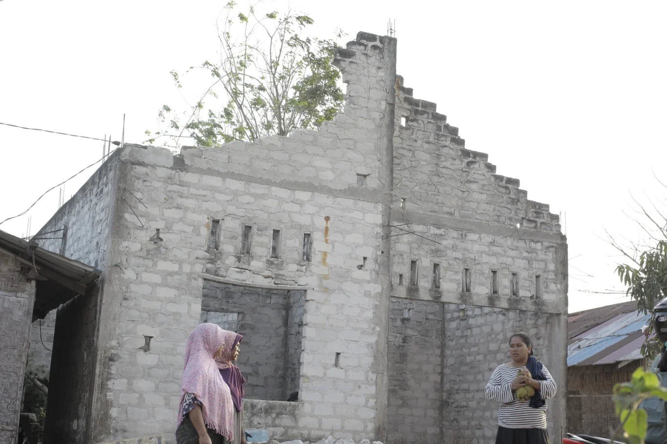 Three women stand before a large, unfinished concrete building with a stepped roof. Two wear headscarves; one holds a green fruit.