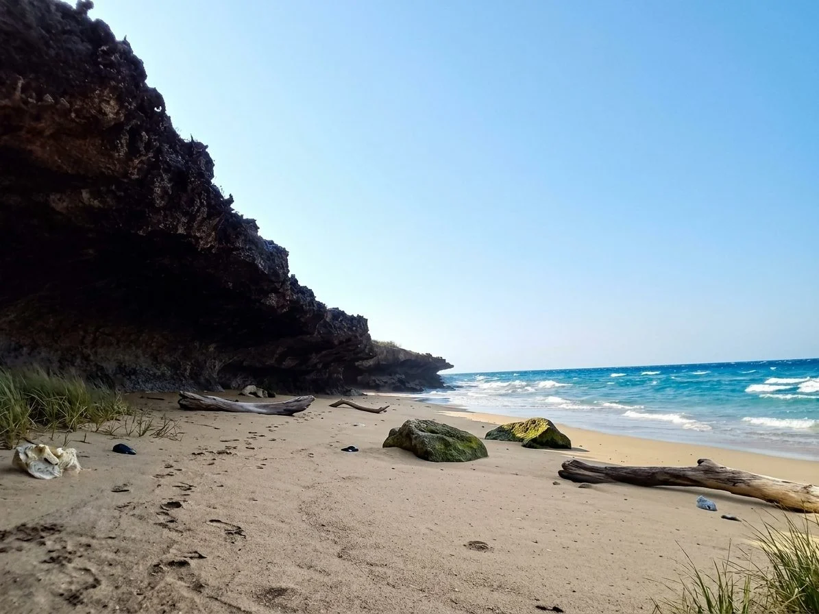 A sandy beach with a dark, overhanging rocky cliff on the left, blue-green ocean waves on the right, and a clear blue sky.