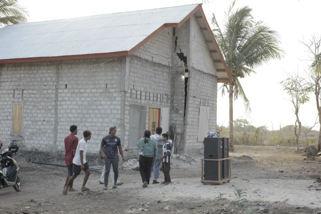 A group of people walks on dry, dusty ground near a grey cinder block building with a large vertical crack, under a bright sky with sparse trees and a palm tree.