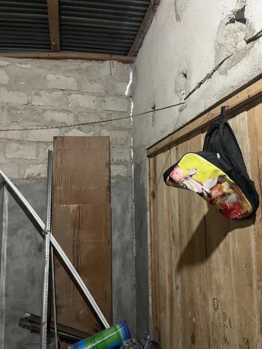 A rustic room corner with concrete block walls, wooden planks, and a corrugated roof, featuring a colorful backpack and light from a wall crack.