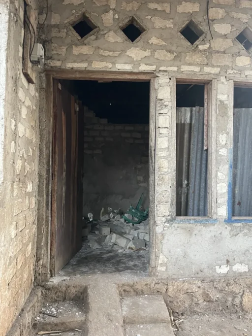 A weathered stone and concrete building facade features a partially open wooden door revealing rubble and a green chair inside. Two window openings are covered with corrugated metal.