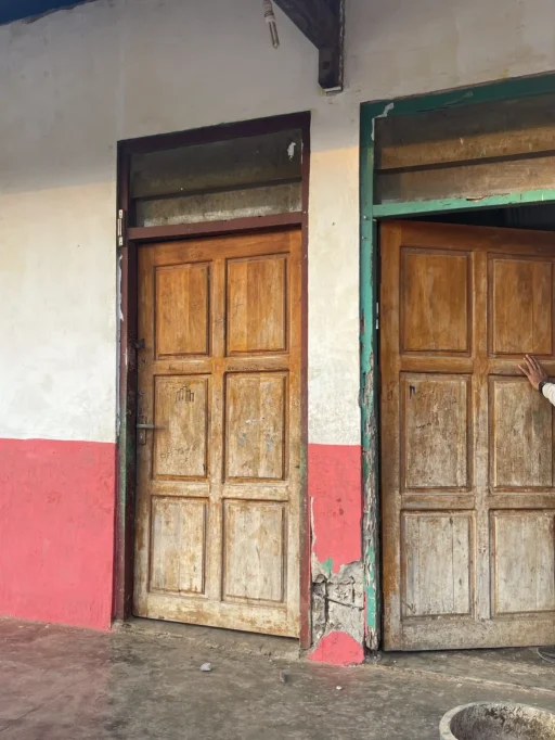 Two weathered wooden doors, one closed and one slightly ajar, are set into a white and red wall with a concrete floor.