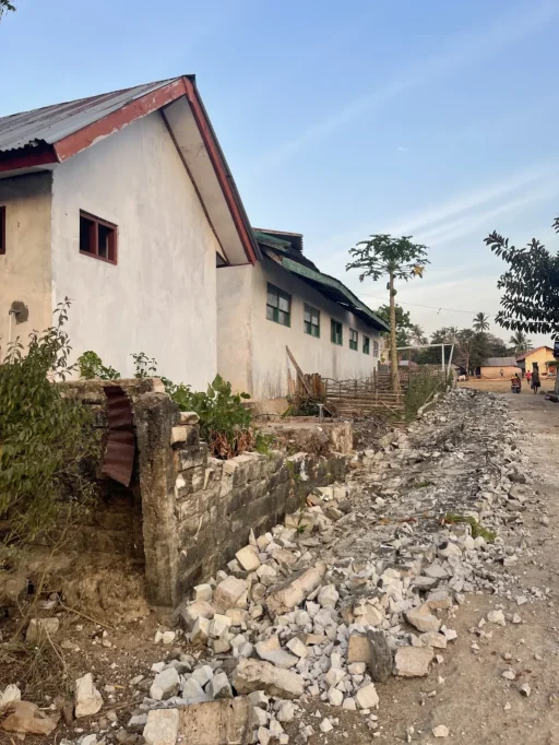 A white building with a red-brown roof stands beside a dirt road covered in concrete rubble under a clear blue sky.